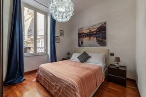 A bedroom with a large bed, a nightstand, a lamp, a painting of a cityscape, and a window with curtains. - A pochi passi da Fontana di Trevi (Roma)