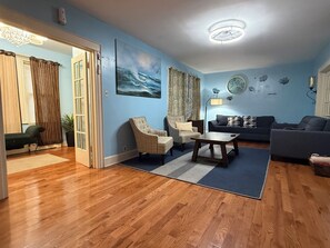 A living room with a wooden floor, a blue wall, a painting, a sofa, a chair, a coffee table, and a lamp. - Spacious Home Prime Location Near Train, JFK, Dining, Shop, Parking & Essentials (Bellmore)
