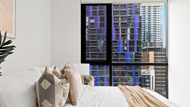 A bedroom with a large window, a bed with patterned pillows, and a view of tall buildings. - Modern 2BR Apt Next to Victoria Market, City Views (Melbourne)