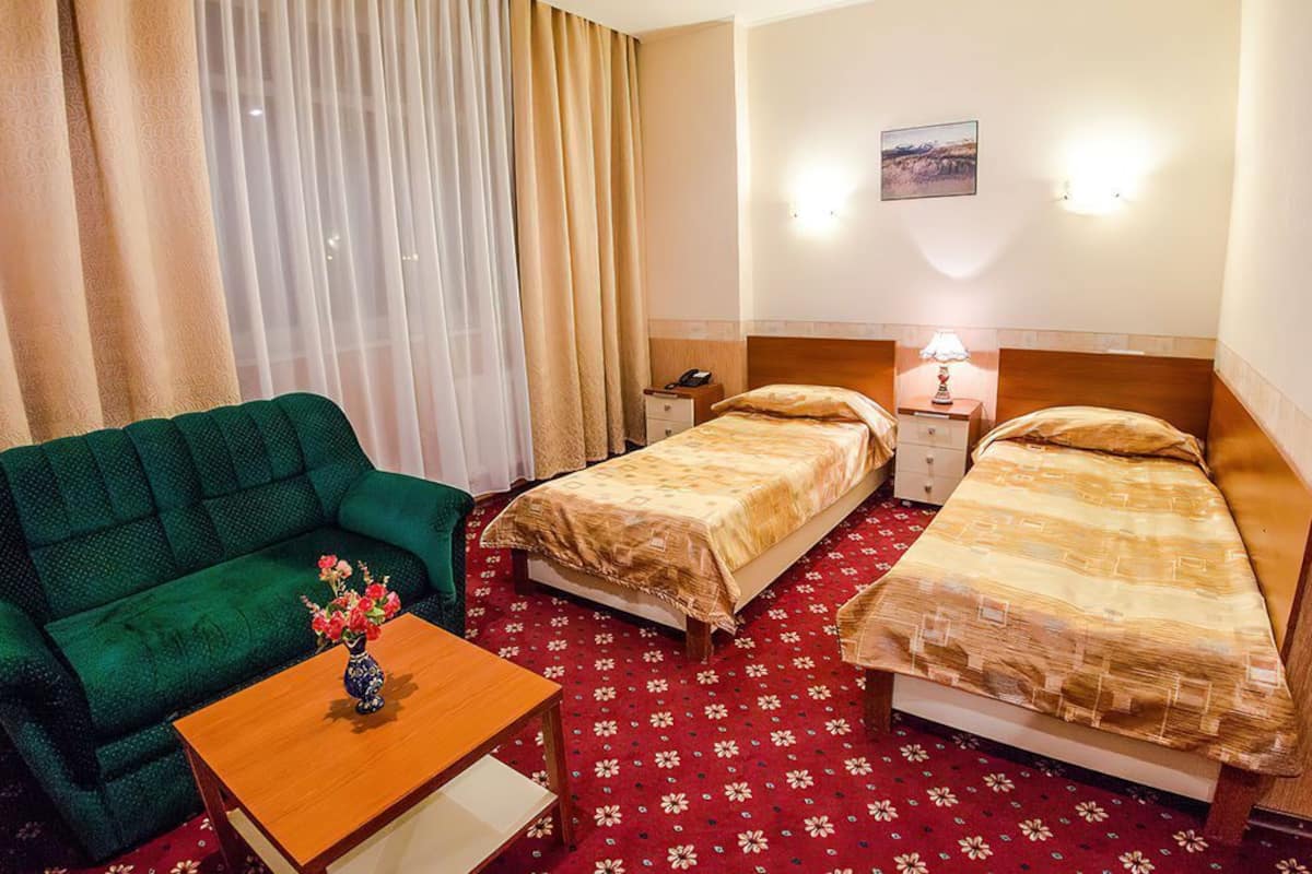 Quarto de hotel com sofá verde, duas camas de solteiro, mesa de madeira com um vaso e carpete vermelho estampado.