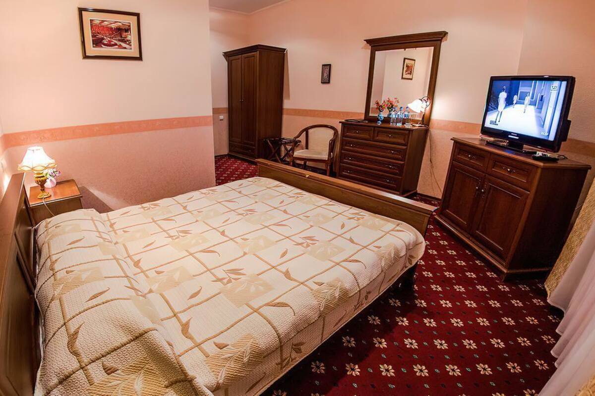 Quarto de hotel com cama, televisão, guarda-roupa, cadeira e secretária com candeeiro.