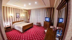 A hotel room with a bed, a TV, a desk with a chair, and a small table with flowers and water bottles. - Reikartz Karaganda (Karagandy)