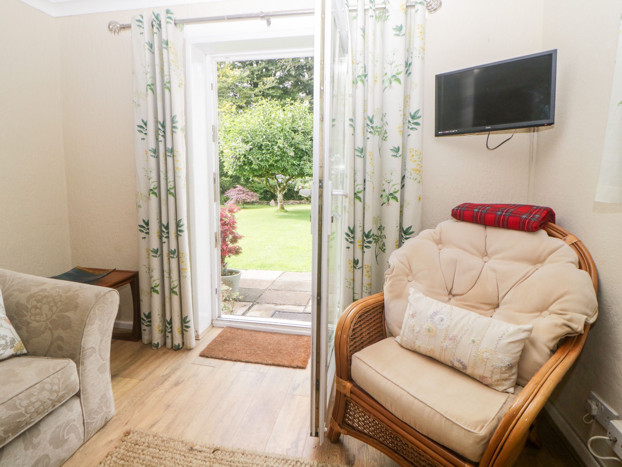 A living room with a sofa, a wicker chair, a television, and a view of a garden through a glass door.