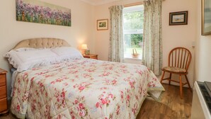 Een slaapkamer met een bloemenbedekker, een gestuufd hoofdeinde, een nachtkastje met een lamp, een raam met gordijnen en een stoel.
