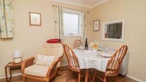 Een eetruimte met een ronde tafel voor vier personen, omringd door rieten stoelen. Er staat een klein bijzettafeltje met een lamp en een ingelijst beeld aan de muur.
