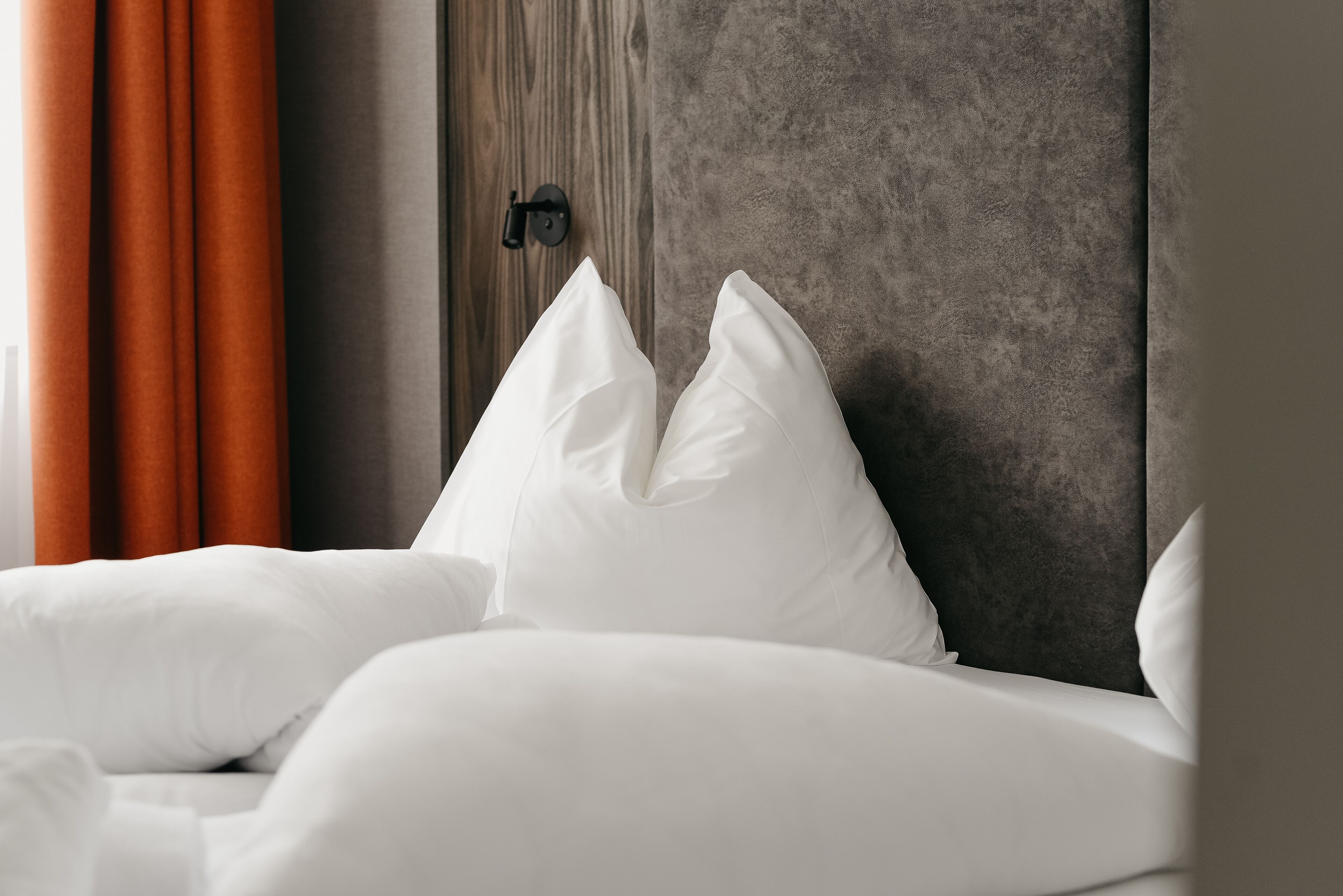 Una cama bien hecha con ropa blanca y almohadas, cabecera de madera y cortinas anaranjadas.