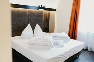 A neatly made bed with white linens and pillows, a wooden headboard, and a window with orange curtains.