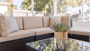 A wicker sofa with beige cushions and patterned pillows, a glass-top coffee table, and a wooden planter with a succulent plant. - Comfortable 3BR | Screened Patio + Grill | WR (Warner Robins)
