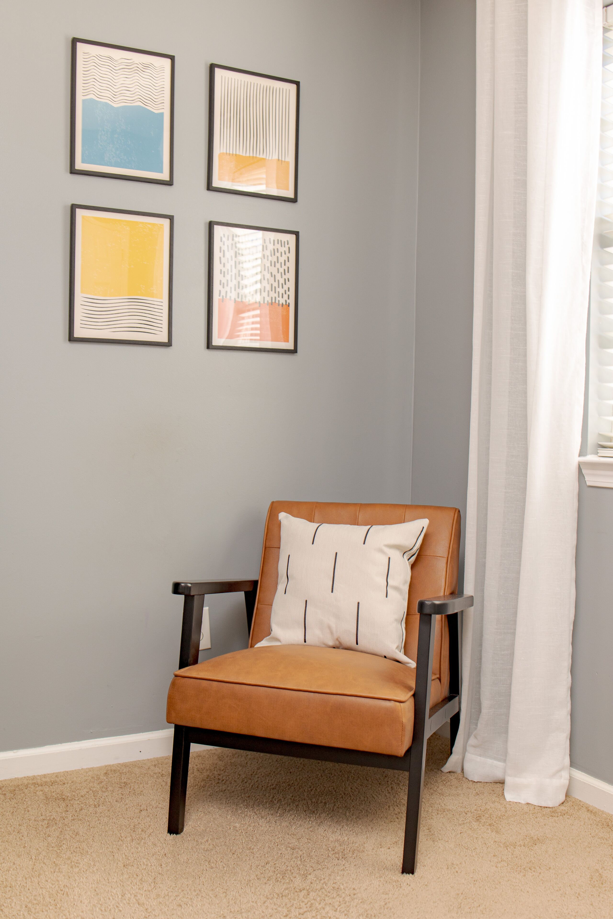 Une chaise avec un coussin, dans une pièce dont un mur est peint en gris clair et où sont accrochées quatre œuvres d’art encadrées.