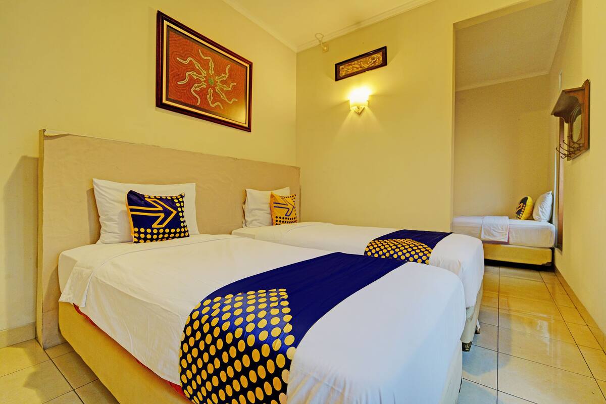 A hotel room with two beds, each with white bedding and blue and yellow polka dot pillows. There are wall-mounted light fixtures and a framed picture above the beds.
