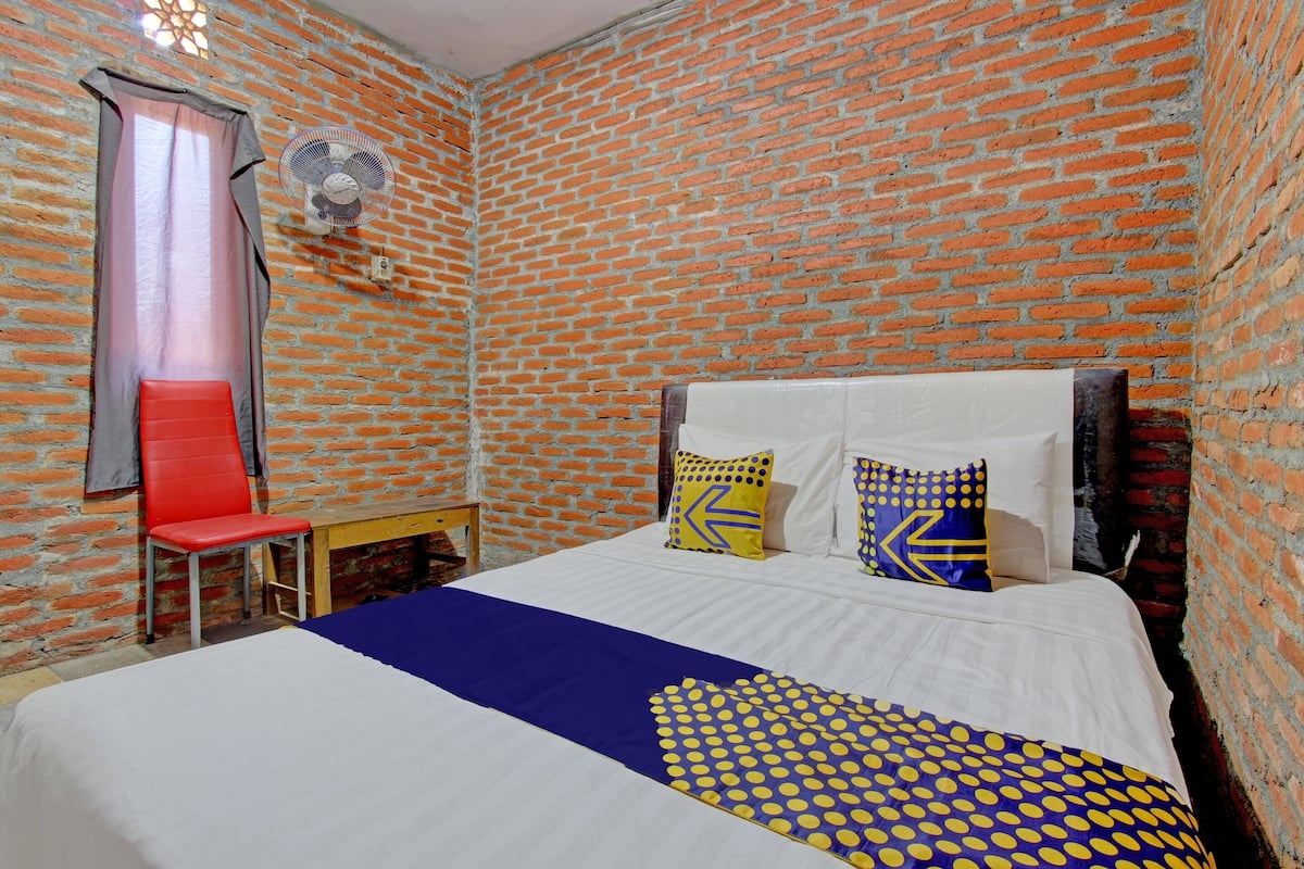 A room with a brick wall, a bed with white bedding and blue and yellow polka dot accents, a red chair, and a fan on the wall.