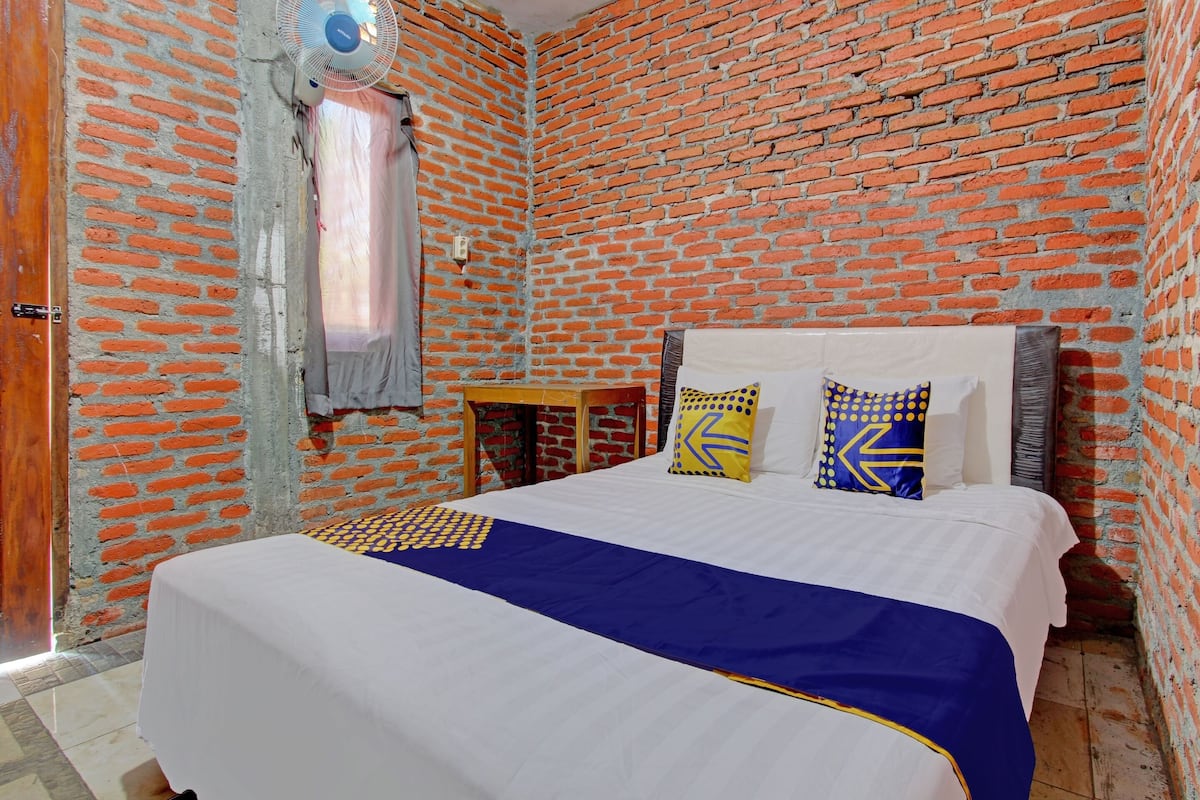 A room with a brick wall, a bed with a white and blue bedspread, and a small table with a fan.