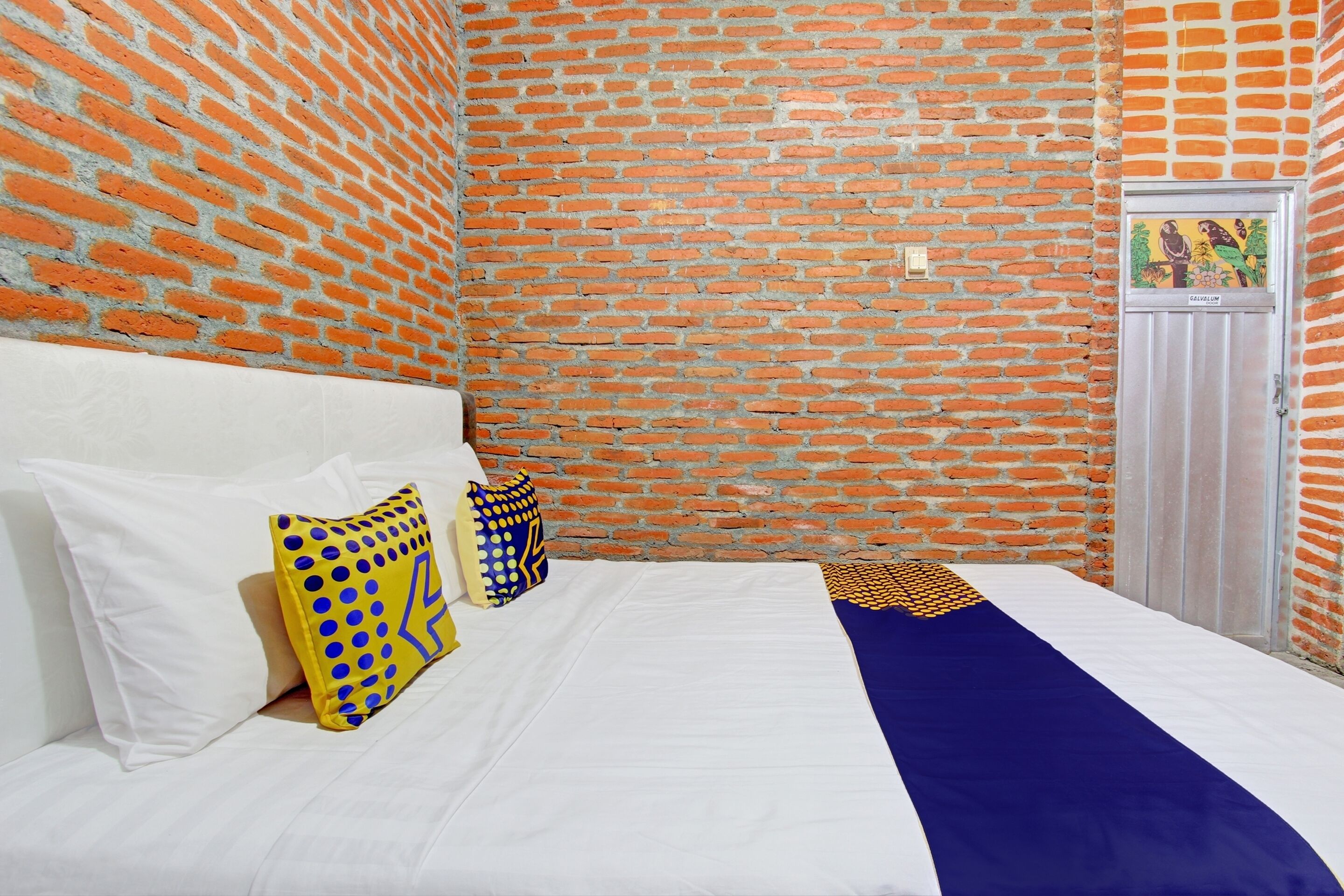 A room with a brick wall, a bed with white bedding and a blue and yellow accent, and a door with a colorful artwork.