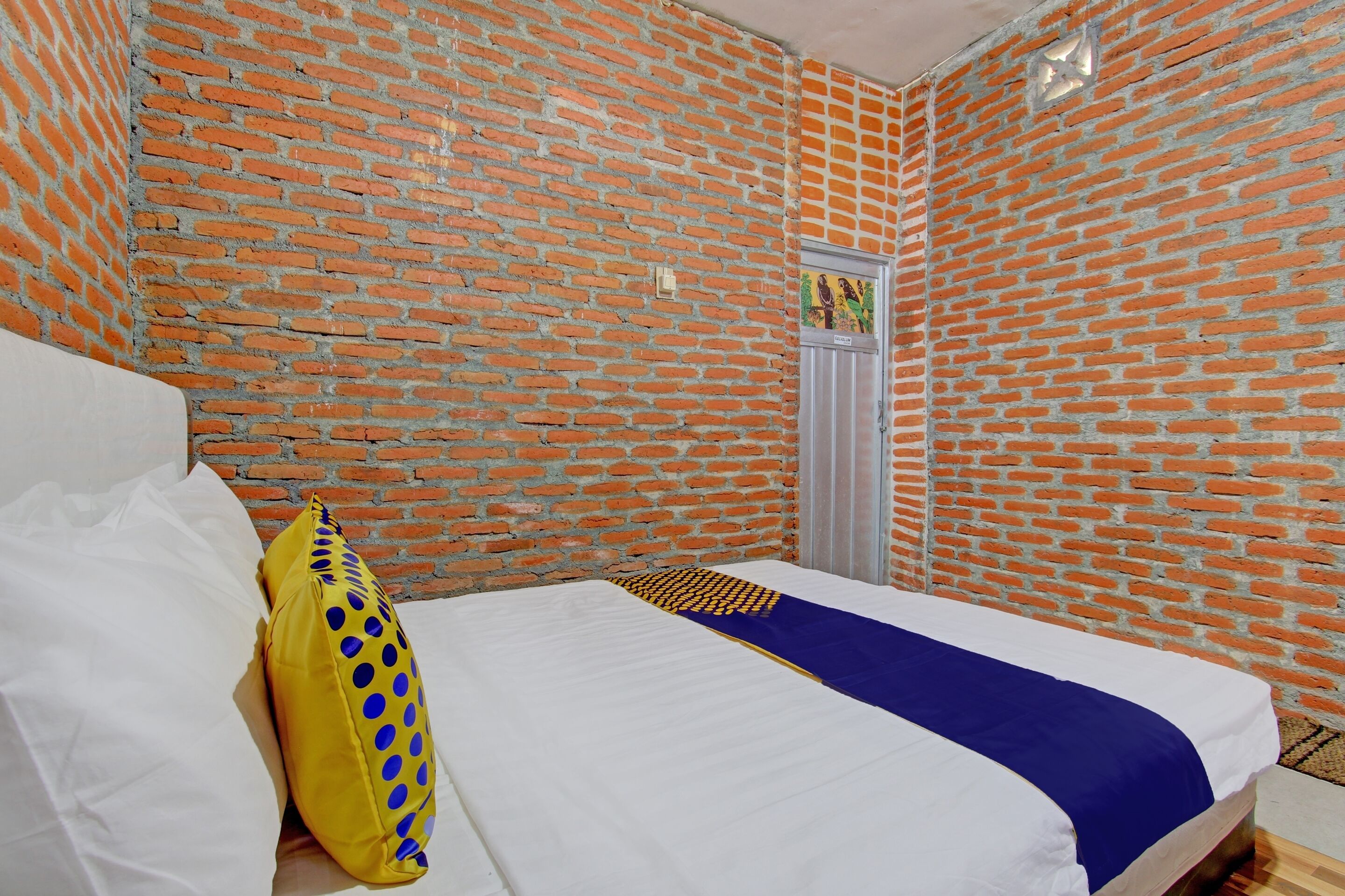 Ein Zimmer mit einer Ziegelwand, einem Bett mit weißer Bettwäsche und einem blauen Akzent sowie einem gelben Kopfkissen mit blauen Punkten.