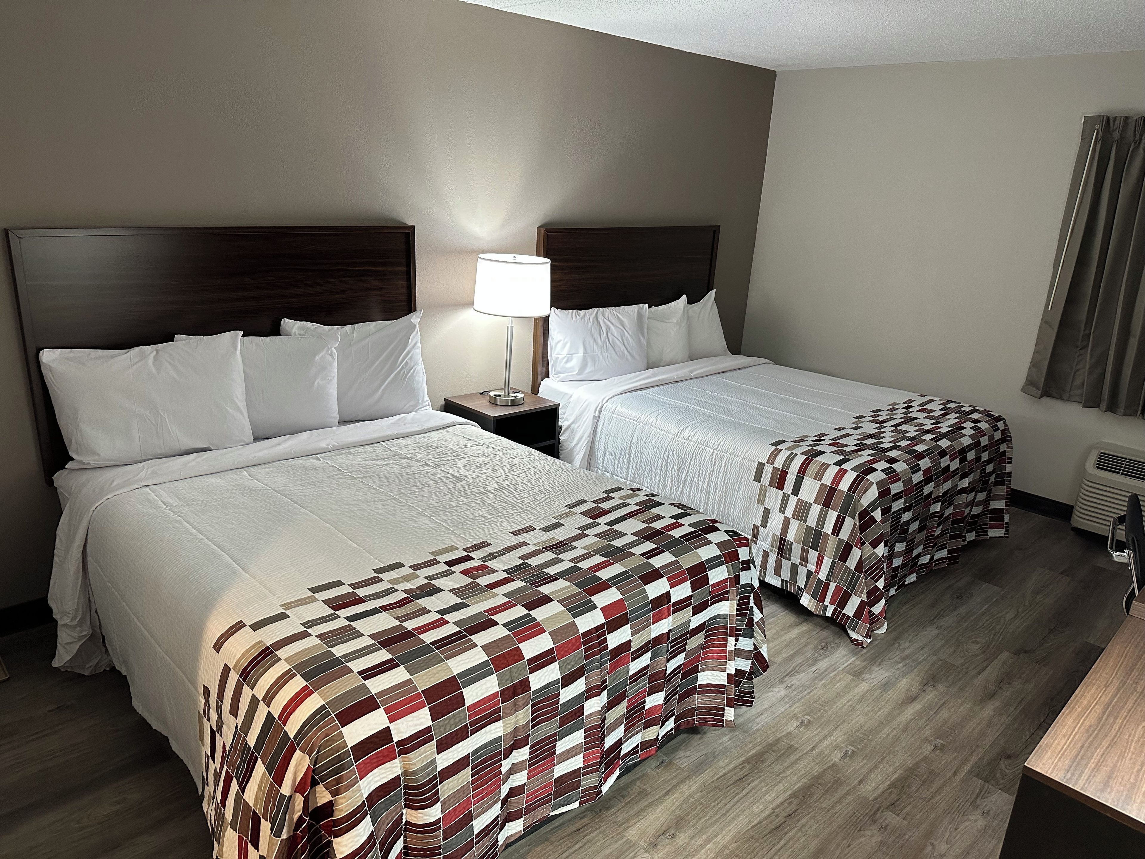 A hotel room with two beds, a nightstand with a lamp, a heater, and a window with curtains.