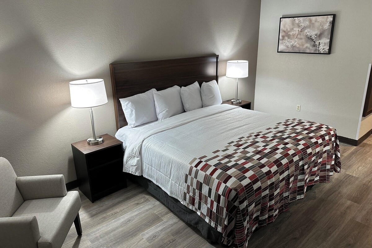 Un letto rifatto con una coperta a quadri, due abat-jour, un comodino, una sedia e un quadro incorniciato appeso al muro.