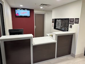 Zona de recepción de un hotel con un televisor mostrando un mensaje de bienvenida, un cartel con los horarios de registro de entrada y salida, y un aviso sobre los artículos de la habitación.
