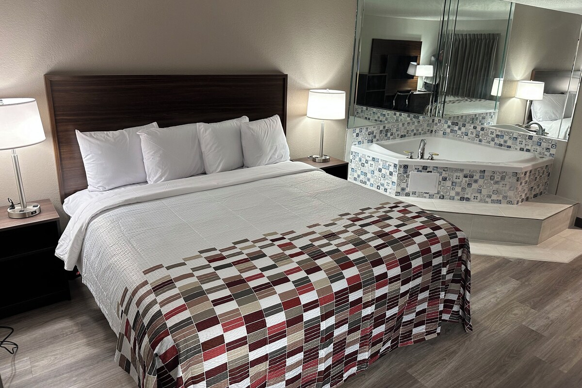 A hotel room with a large bed, a bedside table with a lamp, a bathroom with a bathtub, and a large mirror.