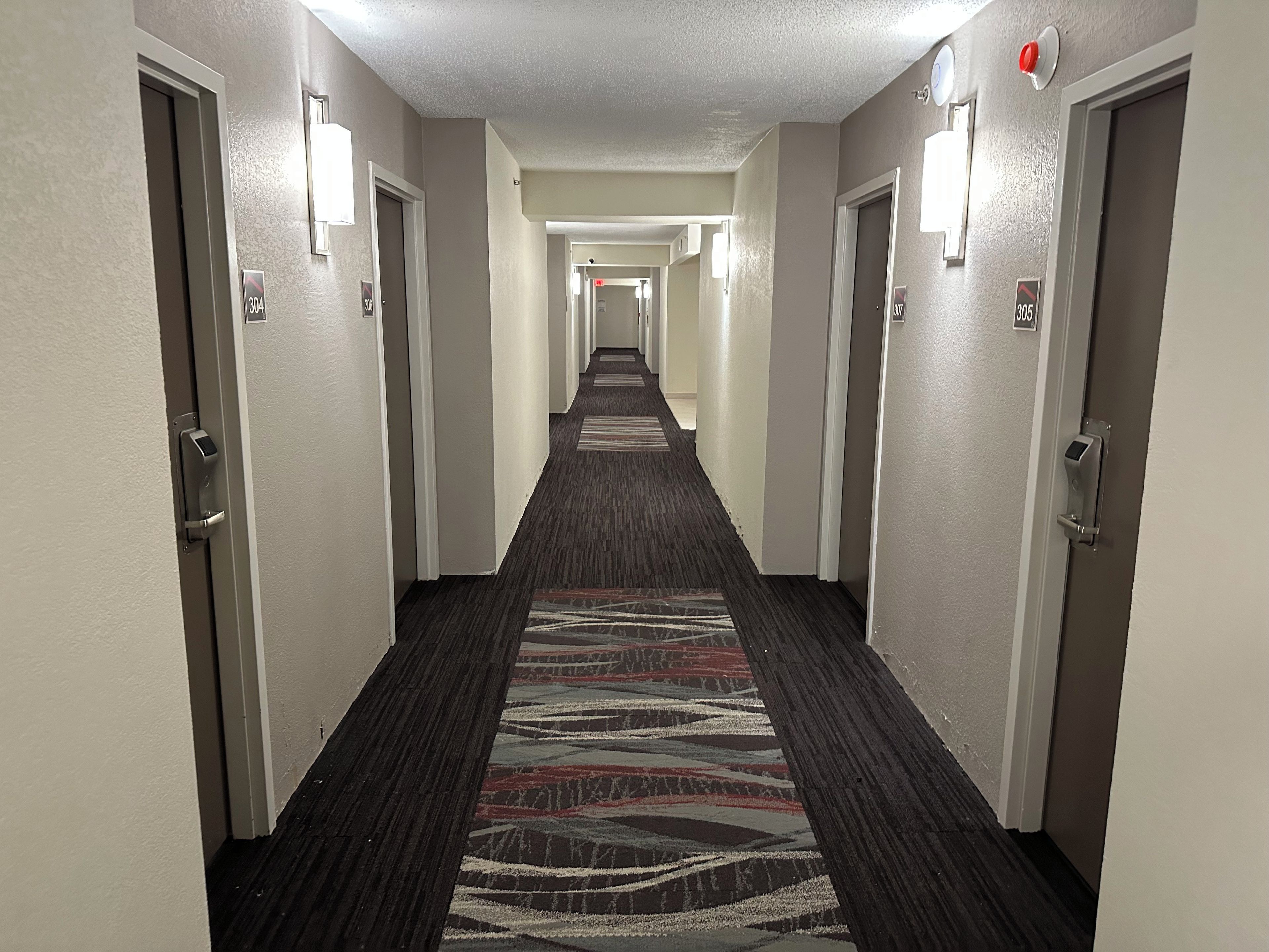 A hotel hallway with carpeted floors, doors on both sides, and wall-mounted light fixtures.