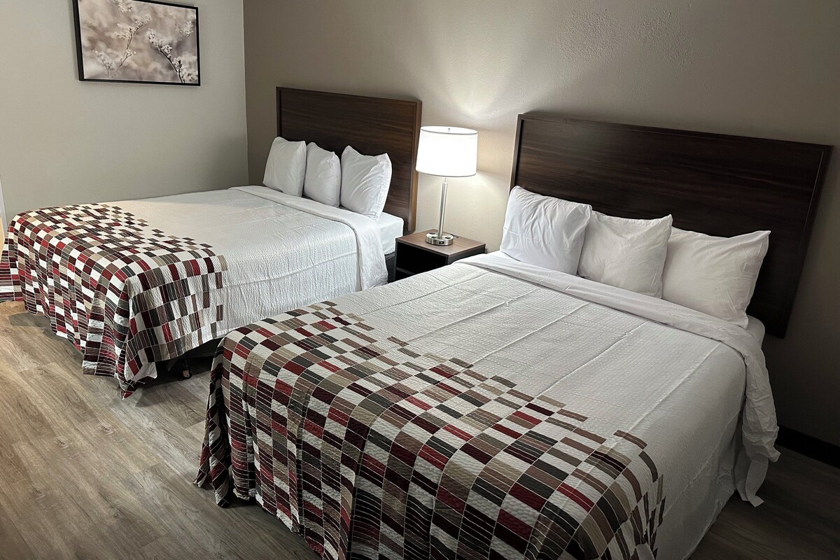 A hotel room with two beds, each with a quilted bedspread, a bedside table with a lamp, and a framed picture on the wall.