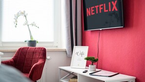 A room with a red wall, a TV mounted on it displaying 'NETFLIX', a red chair, a white desk with a framed 'Wifi' sign, a small potted plant, and a window with white curtains.