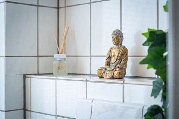 Une salle de bain aux carreaux blancs, une statue de Bouddha dorée, un petit récipient blanc contenant des tiges de roseau et une plante verte.