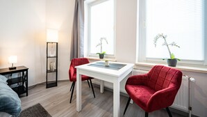 A modern bedroom with a bed, a bedside table with a lamp, a white desk with a chair, and two red chairs. There are two windows with blinds and potted plants on the windowsill.