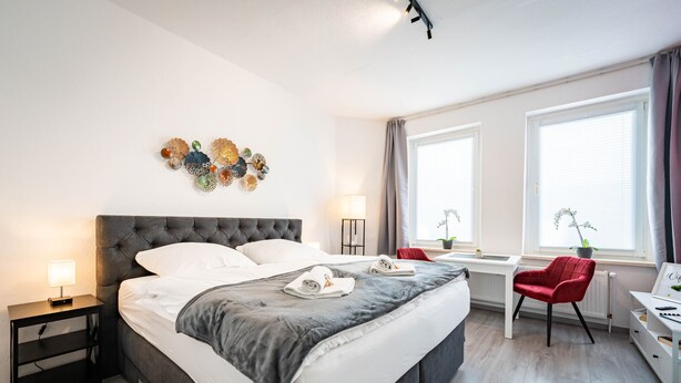 A modern hotel room with a large bed, bedside tables, a desk, and a red chair.