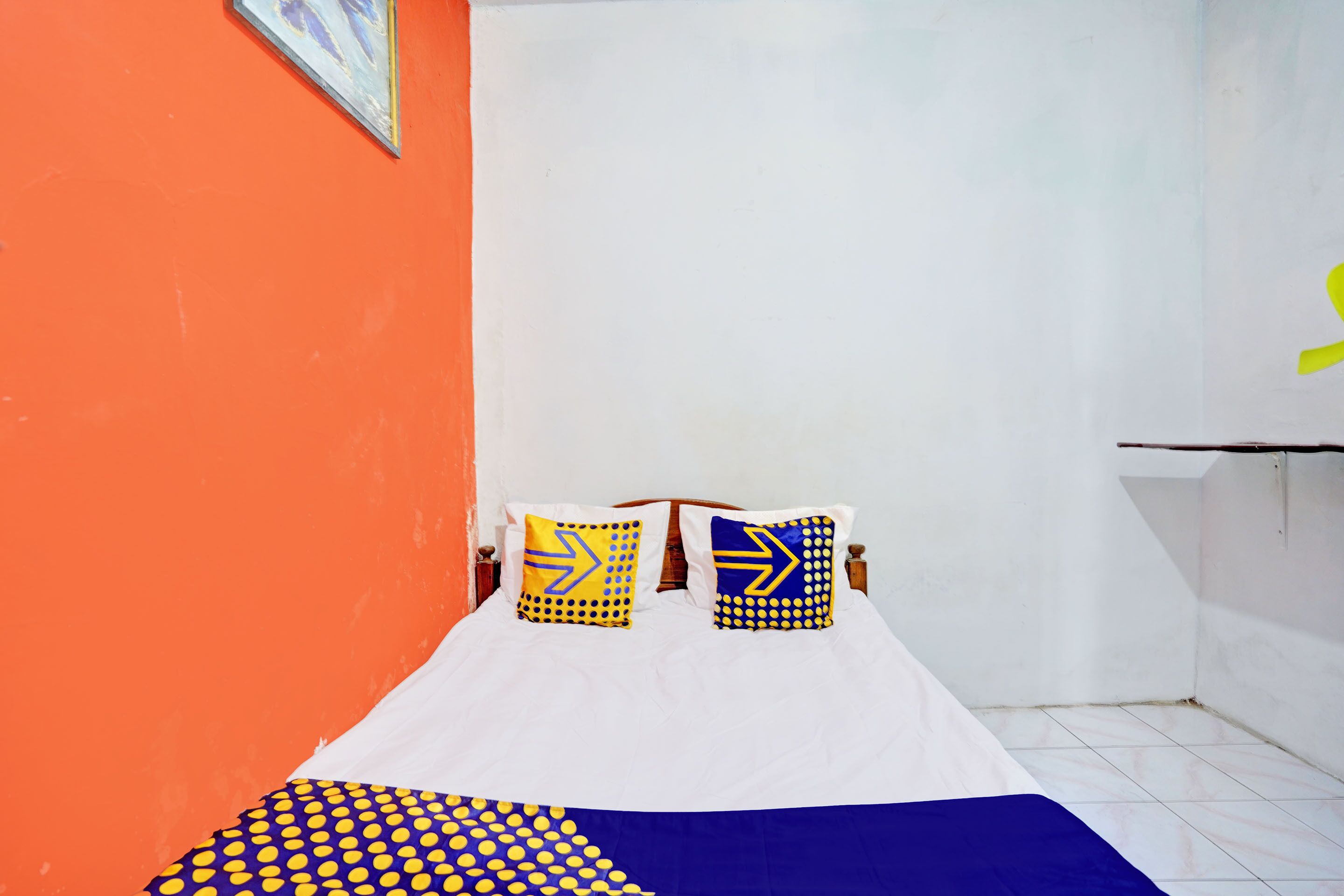 A bed with white bedding and two pillows with yellow and blue patterns, a blue blanket, and a white wall with a painting.