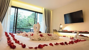 Habitación de hotel con una cama decorada con pétalos de rosa y toallas con forma de cisne, un televisor montado en la pared y vistas a la vegetación por la ventana.