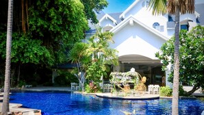 A swimming pool with a curved edge, surrounded by palm trees and a white building with a curved roof. - Pooh Beach Resort & Spa (Pattaya)