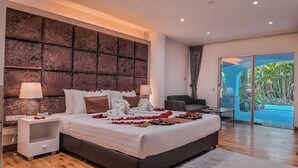 A modern hotel room with a large bed, two bedside tables, a sofa, and a view of a tropical garden through a sliding glass door. - Pooh Beach Resort & Spa (Pattaya)