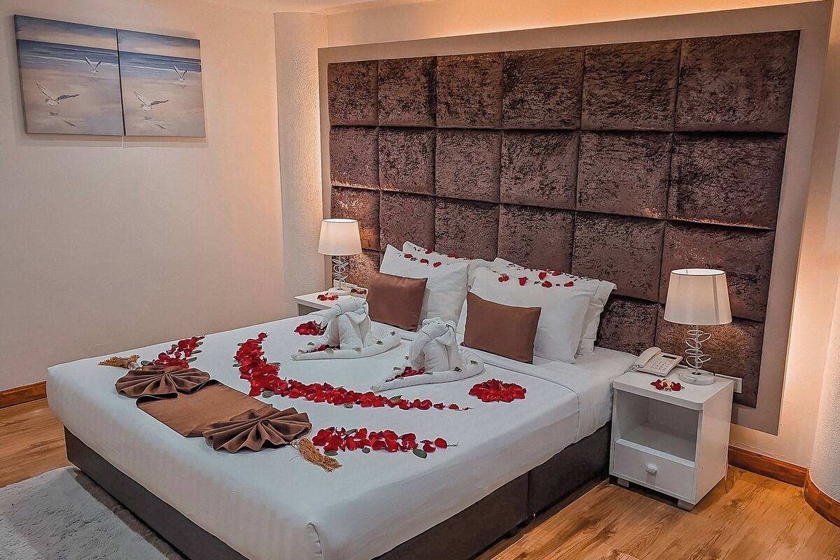 A neatly made bed with rose petals, a bedside lamp, and a wall-mounted artwork.