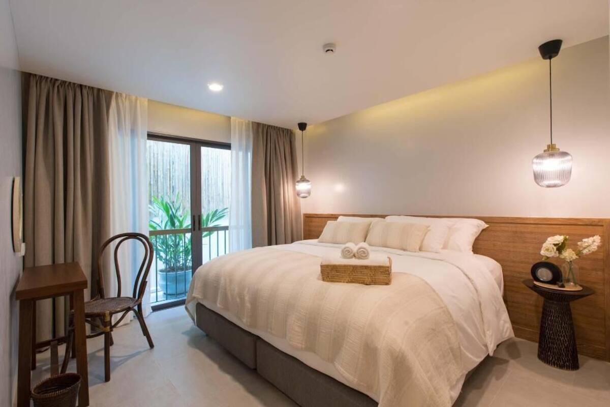 Un dormitorio con cama, una silla, una mesita pequeña, una mesita de noche con flores y vistas al exterior.
