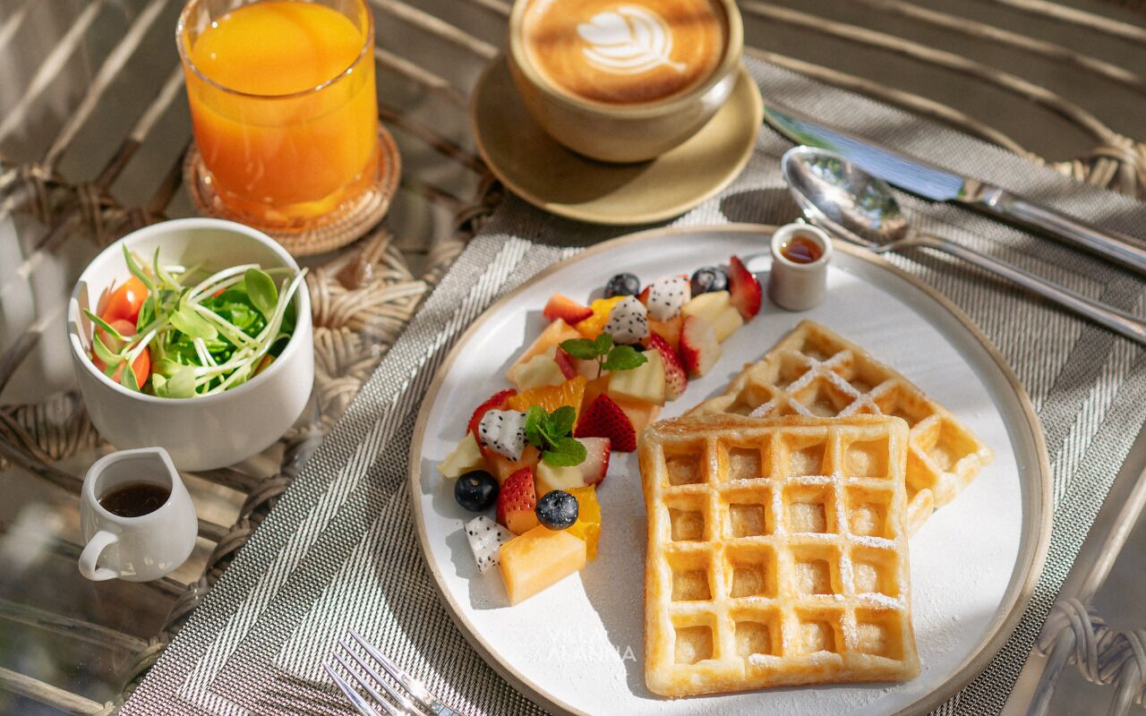 Una escena de desayuno con un plato de gofres, una ensalada de frutas, una taza de café y un vaso de jugo de naranja sobre un mantel tejido.