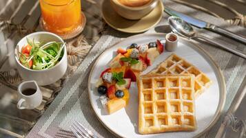 Una escena de desayuno con un plato de gofres, una ensalada de frutas, una taza de café y un vaso de jugo de naranja sobre un mantel tejido.