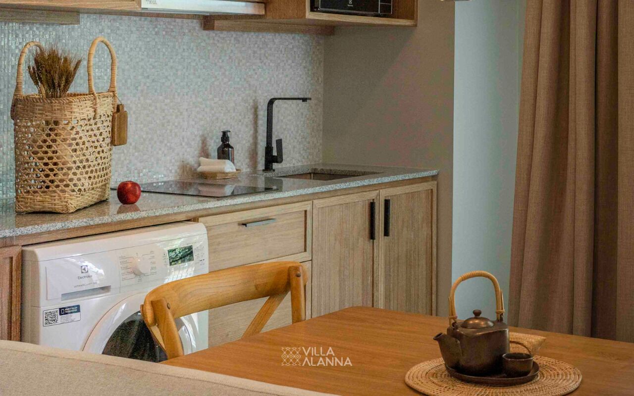 Een compacte keuken met een wasmachine, een houten tafel met een theepot en een rieten mand met gedroogde planten.
