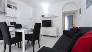 En moderne, minimalistisk stue med en sort sofa, et spiseområde med et hvidt bord og stole, et fladskærms-tv og et køkken med hvide skabe og en mikrobølgeovn.