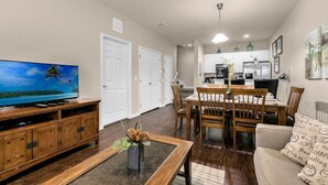 Un salon avec un téléviseur, un meuble de rangement en bois, une table à manger avec des chaises, et un coin cuisine à l’arrière-plan.