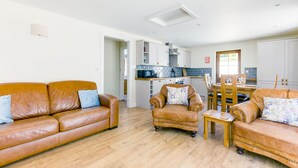 A spacious living area with a brown leather sofa, a small armchair, and a wooden coffee table. A kitchen with white cabinets and a tiled backsplash is visible in the background. - Chy Pol (Wadebridge)