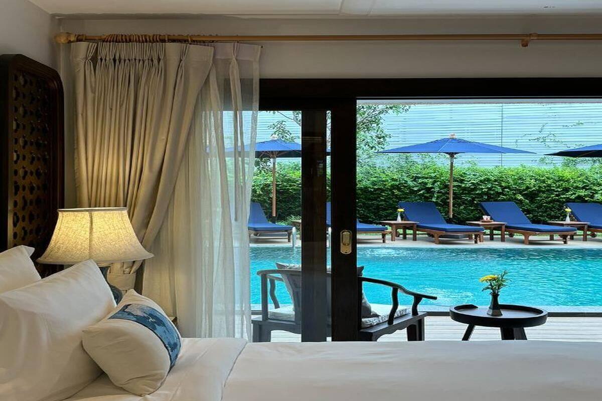 Ein Hotelzimmer mit Bett, Lampe und Blick auf einen Pool mit Liegestühlen und Sonnenschirmen.