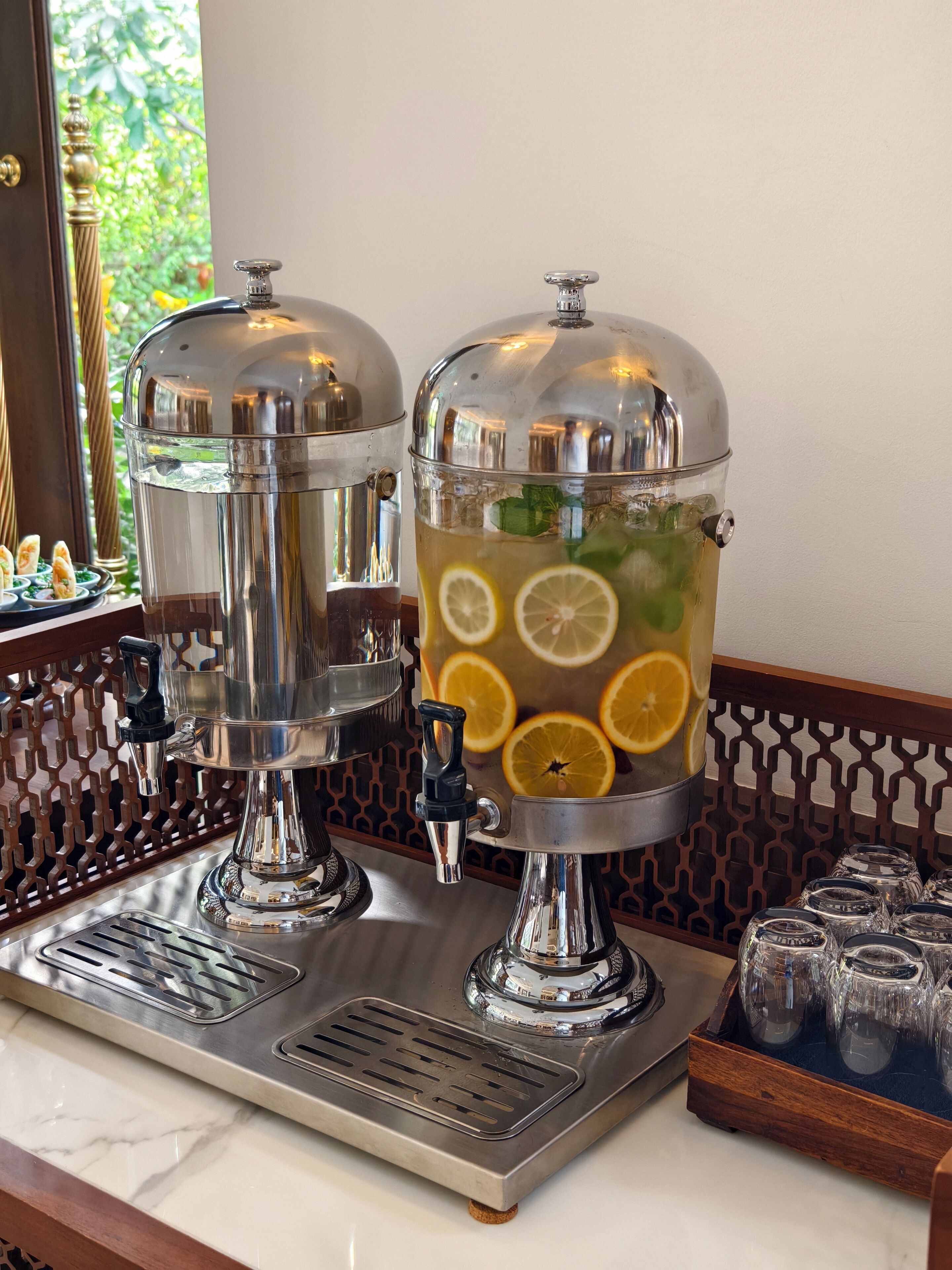 A bar area with two stainless steel dispensers, a tray with glasses, and a window with greenery visible.