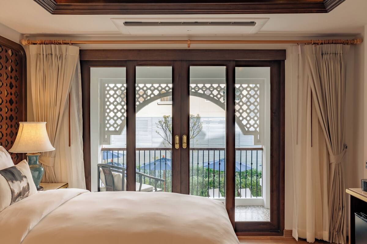 Ein Hotelzimmer mit einem großen Bett, einem Nachttisch mit Lampe und Blick auf einen Balkon mit Gartenmöbeln.