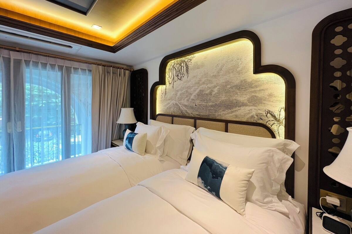 Ein Hotelzimmer mit einem großen Bett, einem Kopfteil mit strukturierter Gestaltung, einem Nachttisch mit Lampe und Blick ins Freie durch durchscheinende Vorhänge.