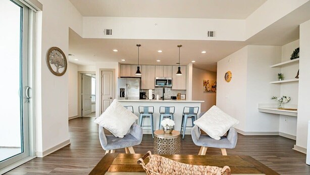A modern living room with a wooden floor, two grey armchairs, a woven basket, and a kitchen in the background.