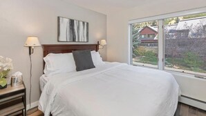 A bedroom with a large bed, two bedside lamps, a nightstand with a lamp, a painting on the wall, and a window with a view of houses. - Awesome Views; Ski In/Out & lift is 100 steps from the Door (Telluride)