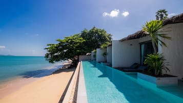 A poolside view with a beach and ocean, a tree-lined pathway, and a building with a thatched roof.