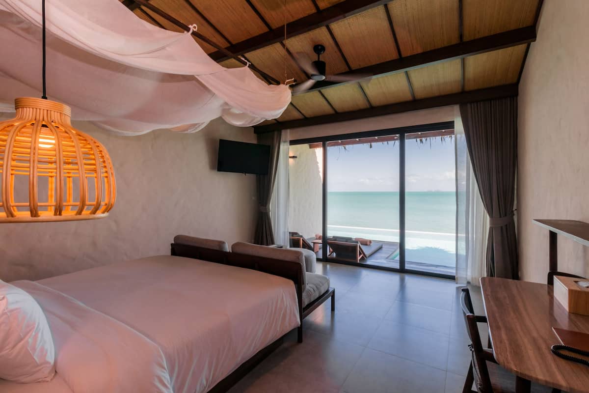 A bedroom with a large bed, a wooden table, a TV mounted on the wall, and a view of the ocean through large windows.