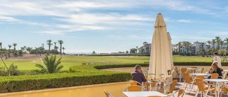 An outdoor dining area with tables and chairs, a large umbrella, and a view of a golf course.