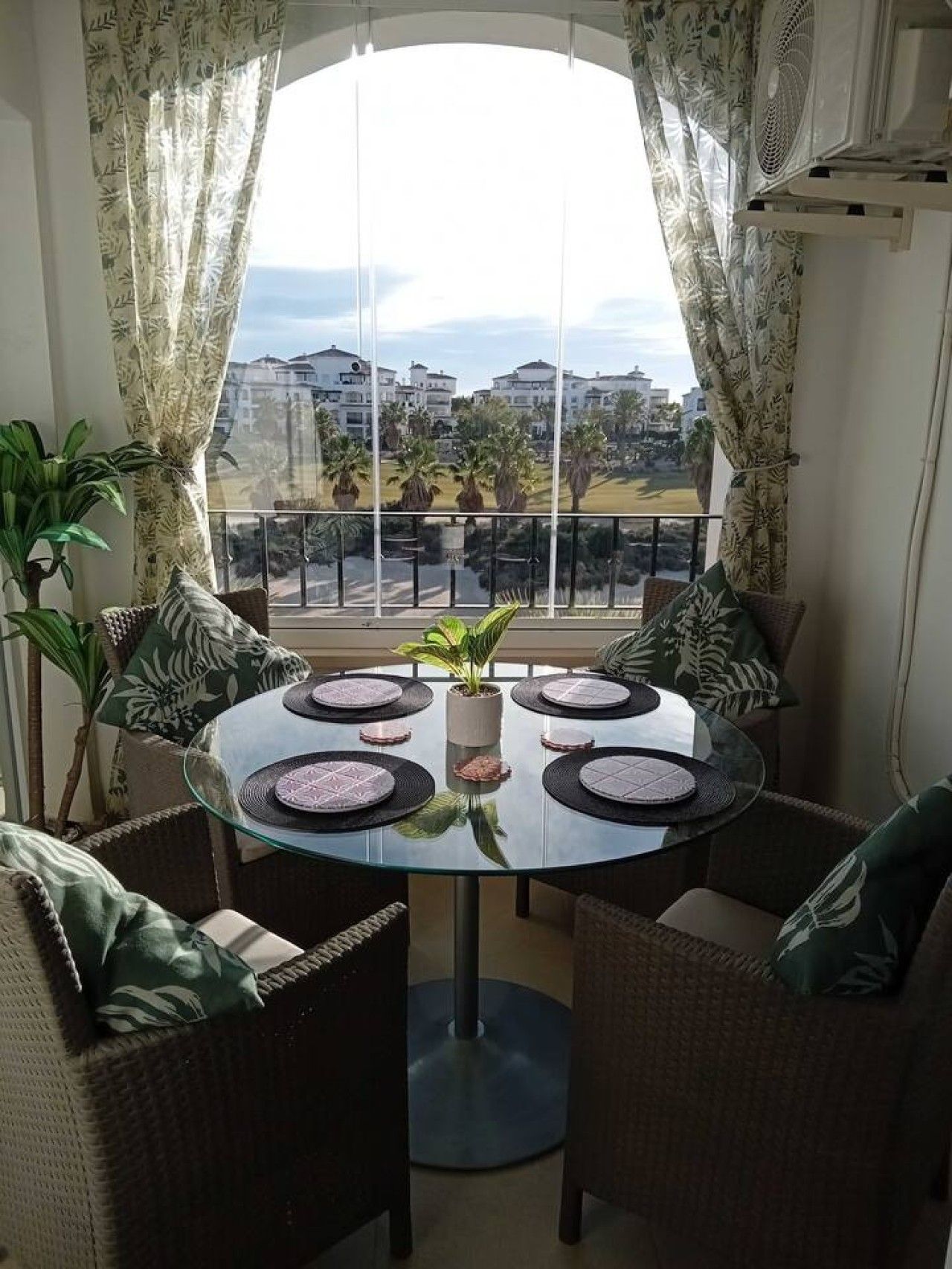 A balcony with a glass table and chairs, a potted plant, and a view of buildings and palm trees.